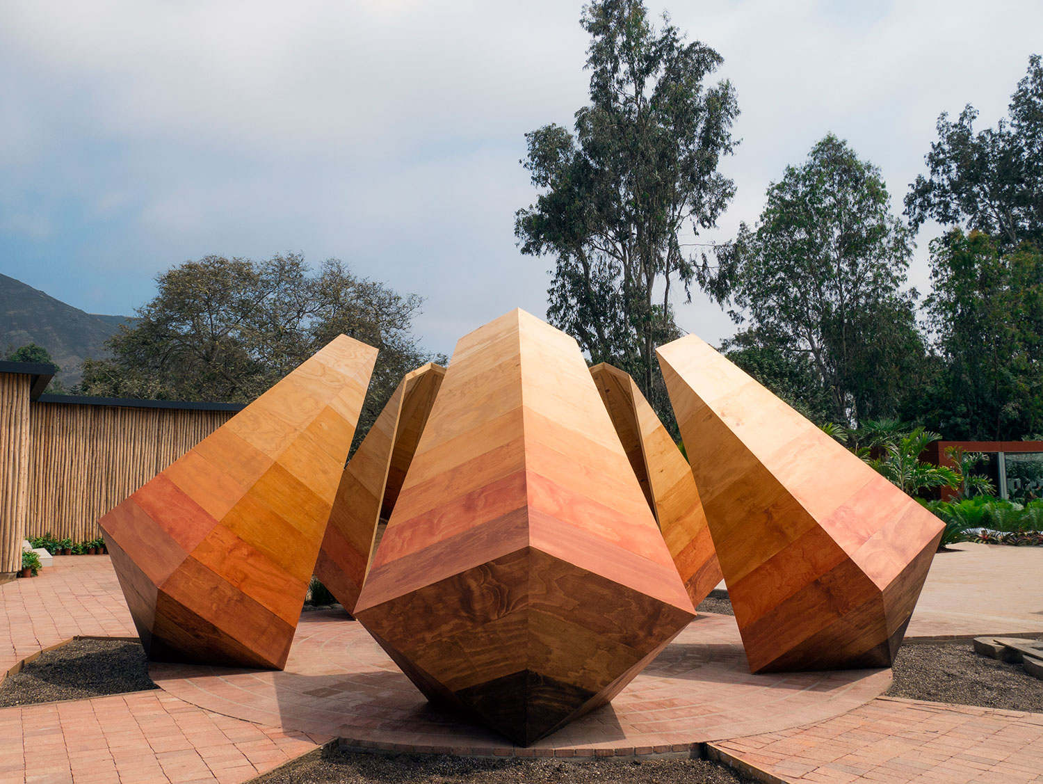 PATIO DEL ENCUENTRO - JUAN SEBASTIÁN RICO. Cinco pirâmides em madeira orbitam em um jardim circular e desafiam a gravidade, unidas pela base. A matéria-prima natural é destacada, para trazer a mensagem de preservação e da importância da certificação da madeira. As esculturas também emitem luzes e sons, criando outras conexões com os visitantes.