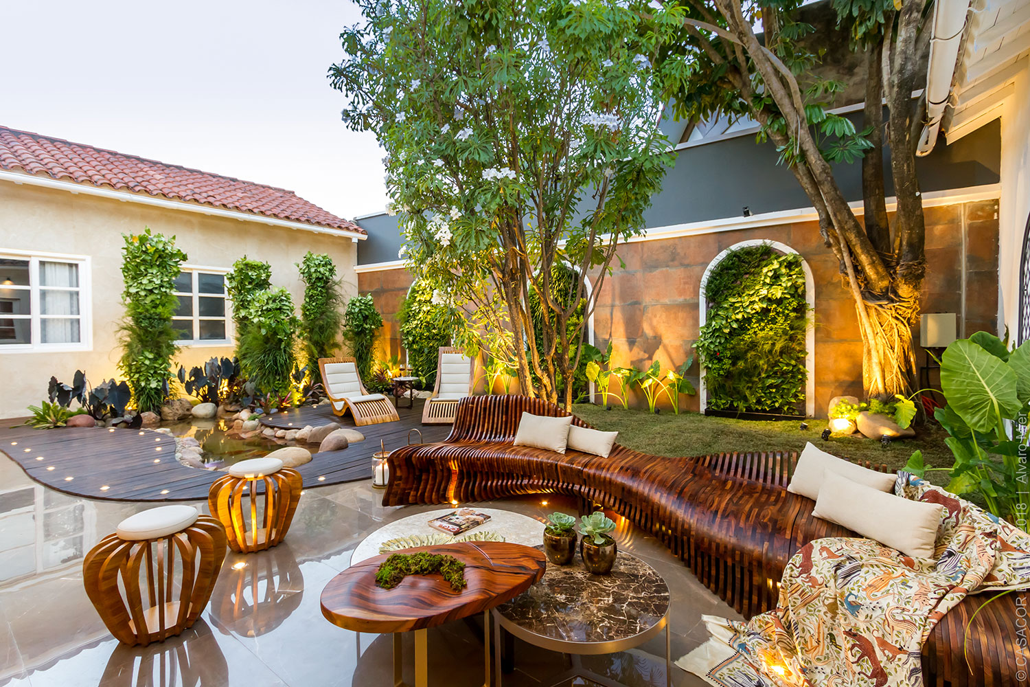 CASACOR Bolívia 2018. El Patio Viviente - Natalia Murillo, Giuseppe Di Domenico E Silvia Oliva. Um jardim vertical surge dos arcos da casa, junto com as colunas verdes. O cenário se completa com o pequeno lago, rodeado por um deck curvilíneo e um banco igualmente sinuoso. Aliás, ele foi produzido com mais de 120 peças de carvalho branco, com o conceito de fabricação digital.