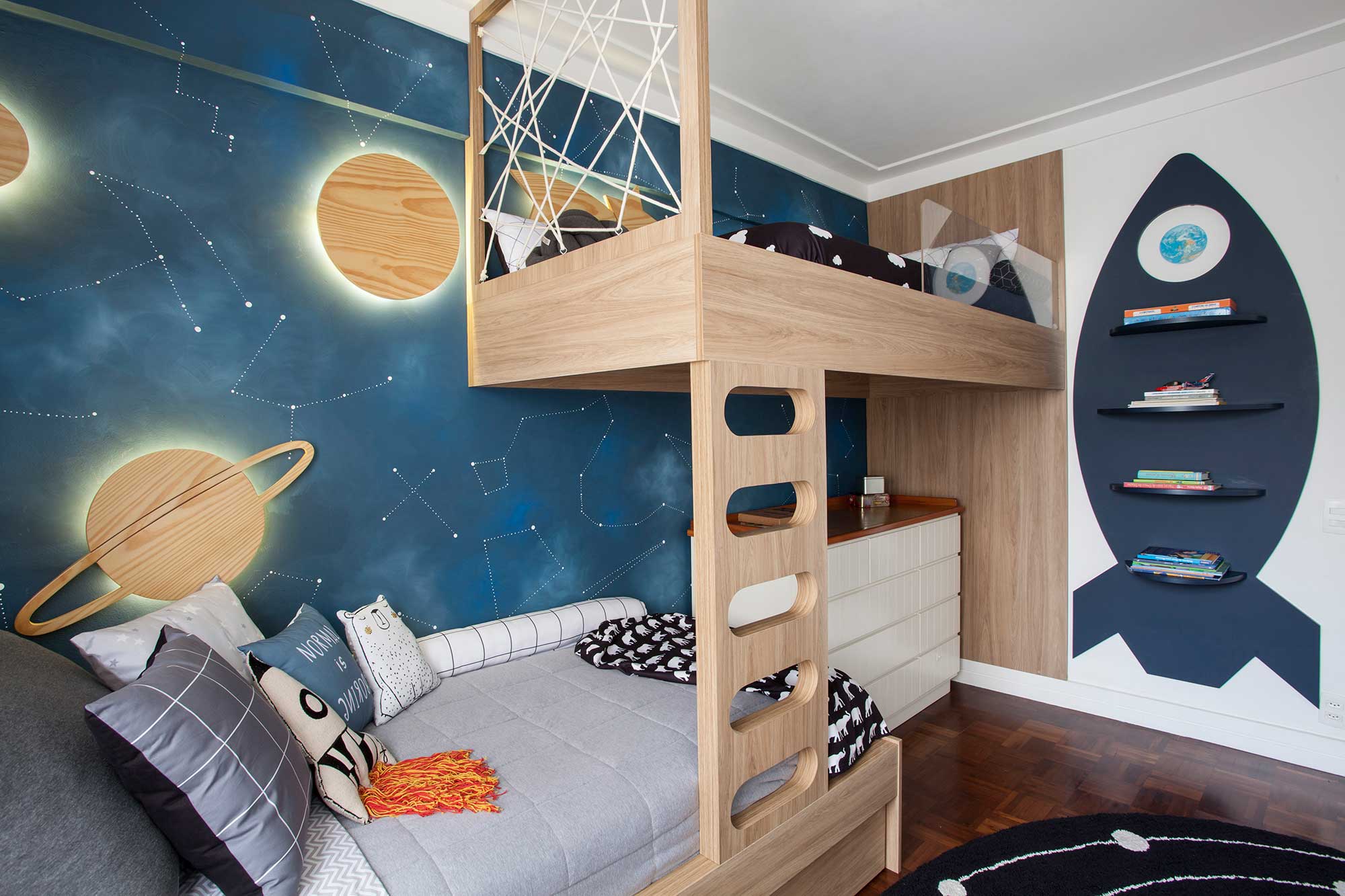 O tema escolhido para agradar dois irmãos é espacial, que determina toda a decoração do quarto. Um dos pedidos era que a decoração estimulasse a criatividade dos pequenos e desse asas às suas criações. Para criar essa atmosfera, as paredes ganharam uma pintura artística com constelações, além de placas de madeira iluminadas por trás em formato de planetas e estrela cadente.