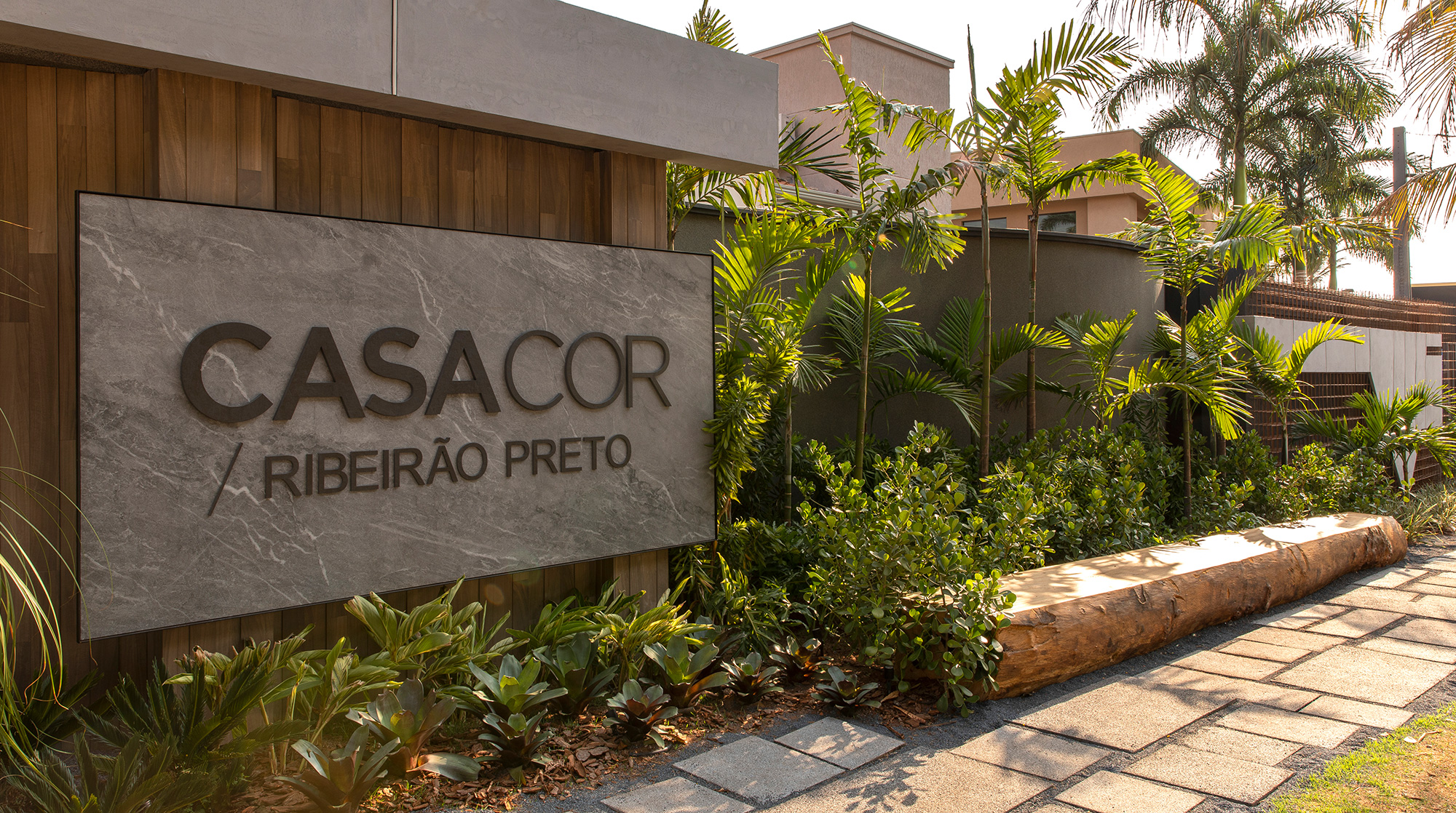 Pajaro Arquitetura - Fachada. O arquiteto e urbanista combinou elementos para receber os visitantes, com sobreposições de diferentes matérias-primas e contrastes entre ângulos e formas, estruturas metálicas e concreto aparente. Entre as referências, aparecem as lastras de porcelanato, as tintas metalizadas, o piso drenante e as texturas acrílicas de pedras naturais. No paisagismo, vasos vietnamitas e um espelho d’água dão equilíbrio ao espaço, que conta também com iluminação de led e som ambiente.