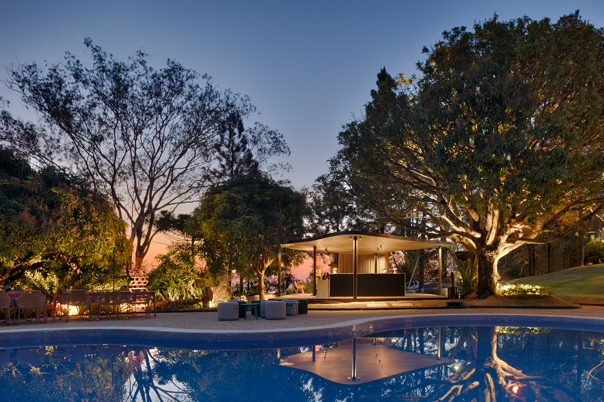 Bar da Piscina - Lucas Lage. Projeto da CASACOR Minas Gerais 2019. O bar de drinks foi inspirado nos rooftops novaiorquinos e sua forma orgânica se molda à piscina. A madeira do deck, de seis centímetros de espessura, emprega a técnica japonesa de queima Shou Sugi Ban. Carbonizada, ganha uma casca resistente ao tempo. O bar em madeira natural ganhou uma frente em pedra retroiluminada e, logo acima, as luminárias de latão escovado trazem pintura dourada em estilo art déco.