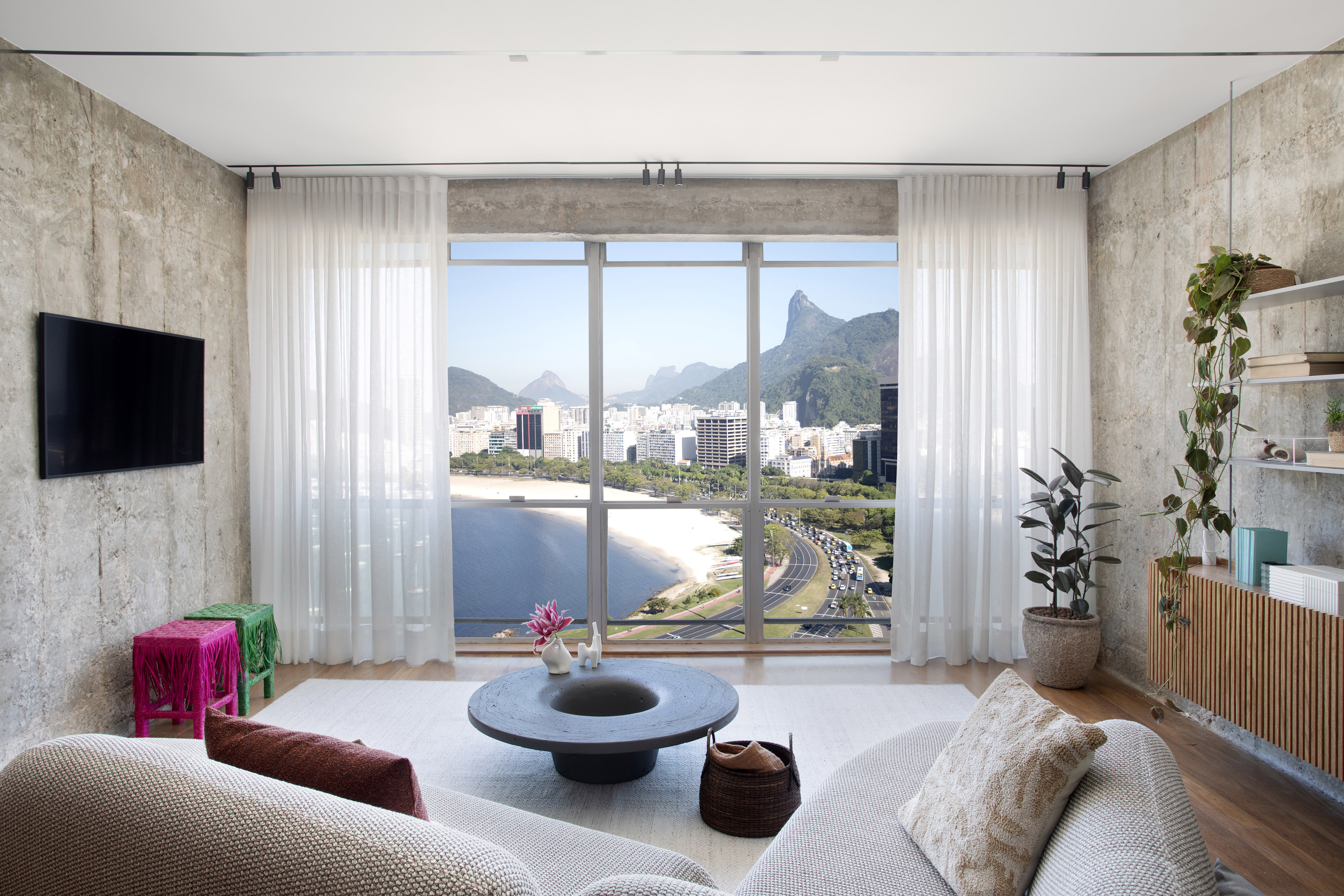 Rafael Ramos emoldura a paisagem carioca neste apartamento de 98 m². Na foto, sala com parede de concreto, vista para a praia e sofá.