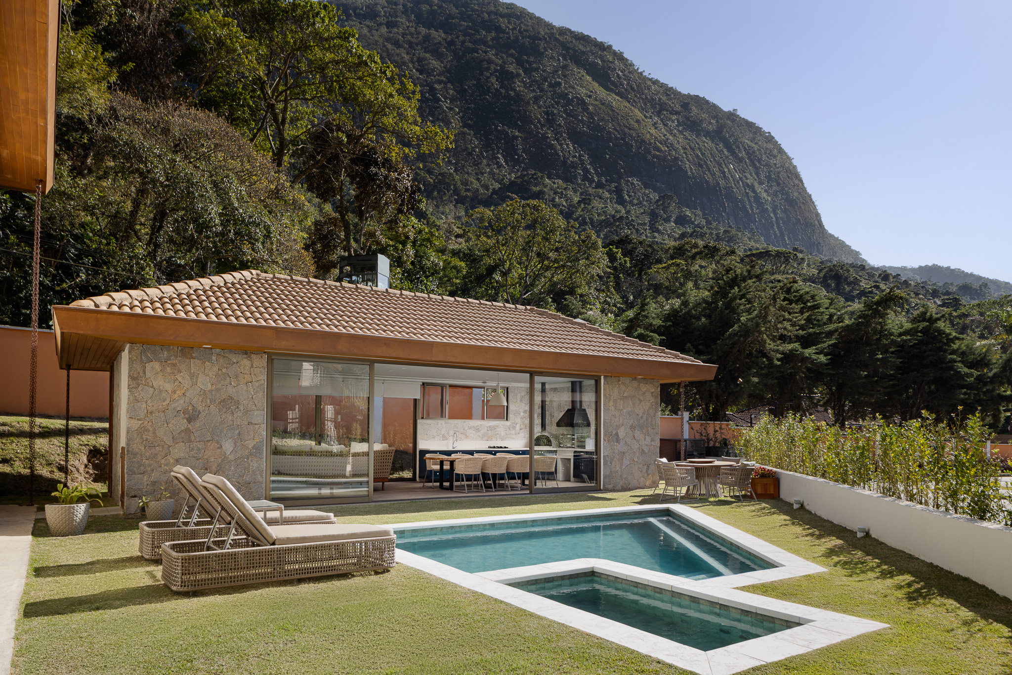 Casa na serra ganha anexo com parede de pedra em projeto de Rafael Mirza. Na foto, área gourmet com churrasqueira e mesa de refeições. Piscina e jardim.